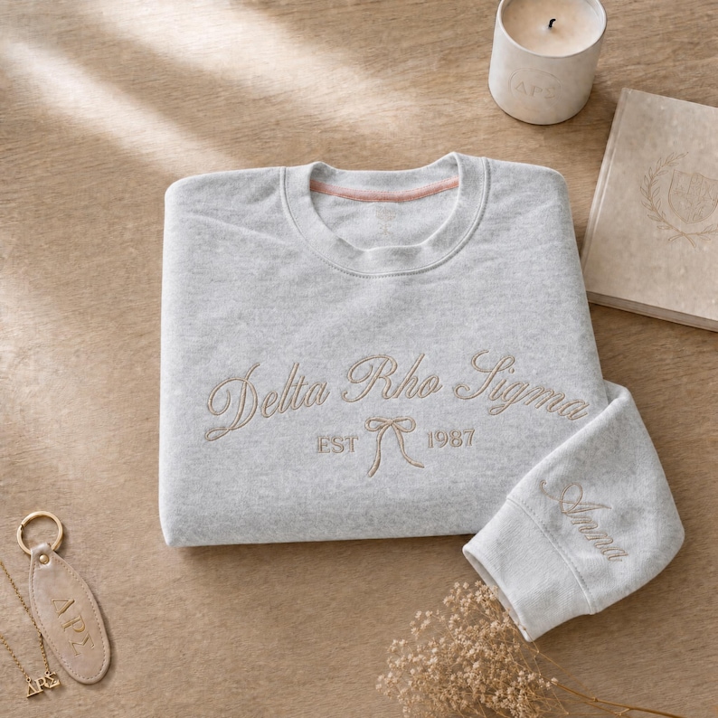 May include: A light gray sweatshirt with the embroidered text "Delta Rho Sigma" and "EST 1987" in a cursive font. The sleeve cuff is embroidered with the name "Anna". A gold keychain and necklace are visible.