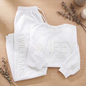May include: A white matching sweat suit set. The sweatshirt says "WIFEY EST 2025" and has the initials "JVM" on the sleeve. The sweatpants say "MRS. WILLIAMS". The set is displayed on a light brown wooden surface.