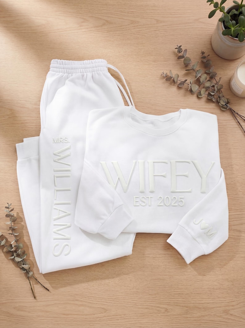 May include: A white matching sweat suit set. The sweatshirt says "WIFEY EST 2025" and has the initials "JVM" on the sleeve. The sweatpants say "MRS. WILLIAMS". The set is displayed on a light brown wooden surface.