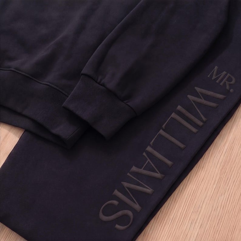 May include: Navy blue sweatshirt and sweatpants set. The sweatpants feature the name "WILLIAMS" vertically down the side, with "MR." above. The set is made of a soft material.