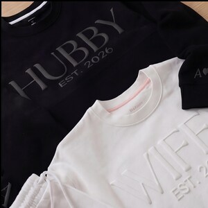 May include: Black sweatshirt with the word "HUBBY" and "EST. 2026" in raised lettering. A white sweatshirt with the word "WIFEY" and "EST. 2026" in raised lettering is also visible. A black beanie with the letter "A" and a heart is also visible.