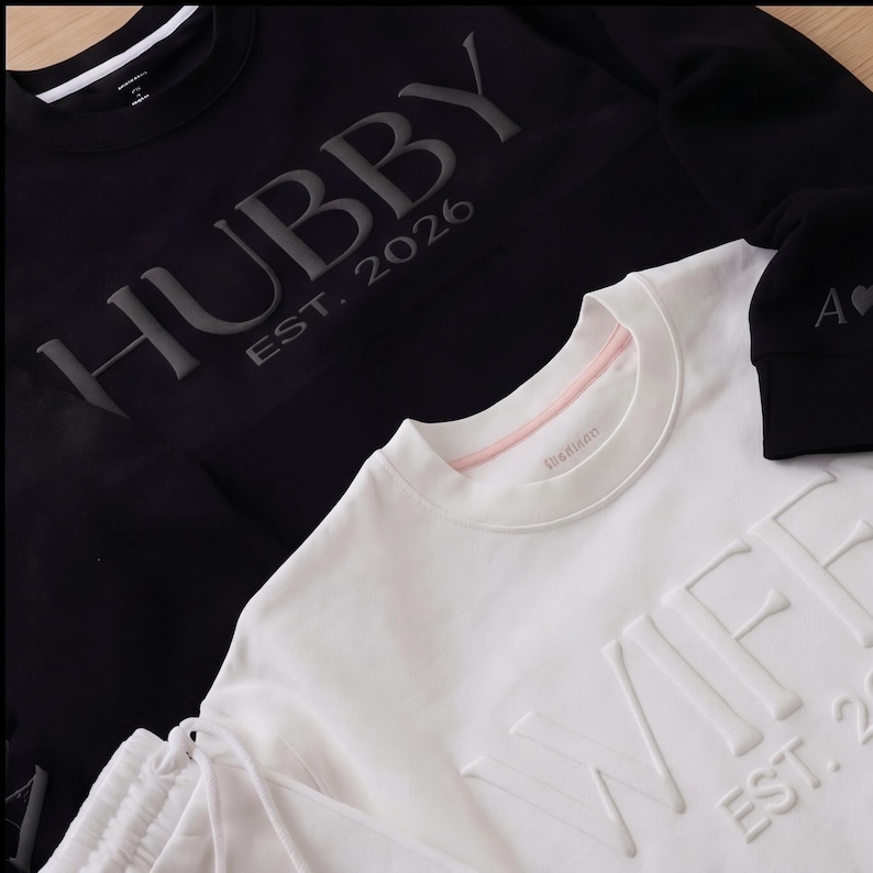 May include: Black sweatshirt with the word "HUBBY" and "EST. 2026" in raised lettering. A white sweatshirt with the word "WIFEY" and "EST. 2026" in raised lettering is also visible. A black beanie with the letter "A" and a heart is also visible.