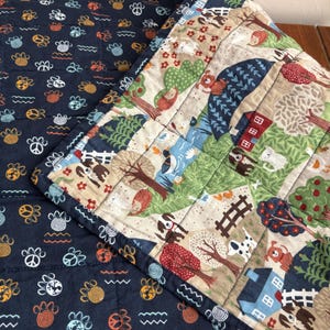 May include: A quilted fabric piece featuring two distinct patterns. One side is dark navy blue with a repeating print of orange paw prints, peace signs, and blue globes. The other side displays a colorful farm scene with houses, trees, and animals.