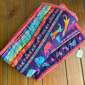 May include: A quilted fabric piece featuring a purple background with colorful cats, fish, and butterflies. The border is a textured coral red fabric. A small white tag attached to the corner displays the text Leaping Cats Red 10x23.