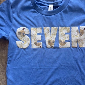 Könnte beinhalten: Ein hellblaues Kurzarm-T-Shirt mit dem Wort SEVEN, das als Applikation auf der Brust angebracht ist. Der cremefarbene Stoff zeigt ein wiederkehrendes Muster aus kleinen gelben Küken und braunen Sprenkeln mit sichtbaren Nähten.