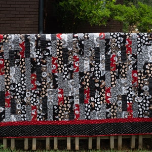 May include: A black, white, and red patchwork quilt with a geometric pattern. The quilt is hanging on a wooden fence.