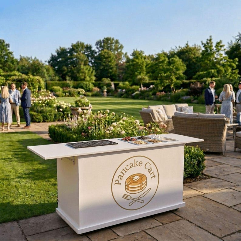 May include: A white pancake cart with a gold logo that reads "Pancake Cart" with a pancake, knife, and fork illustration. The cart has a serving area with food and a black tray. The cart is outdoors on a patio.