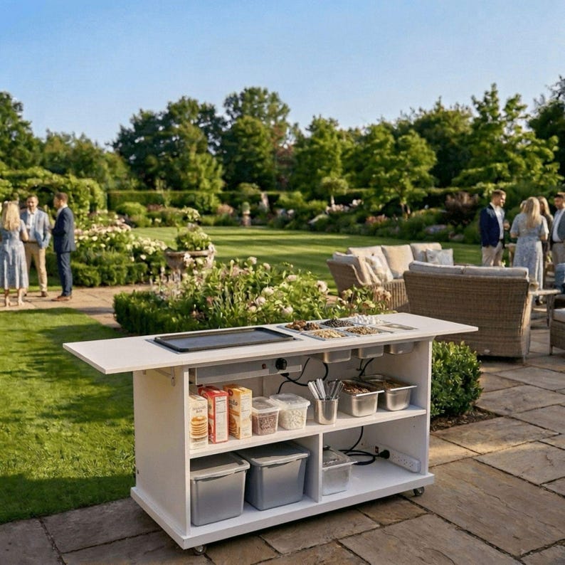 May include: A white outdoor serving cart with a black griddle and food containers. The cart has shelves with food items and storage bins. It is set up on a stone patio with a green lawn and people in the background.