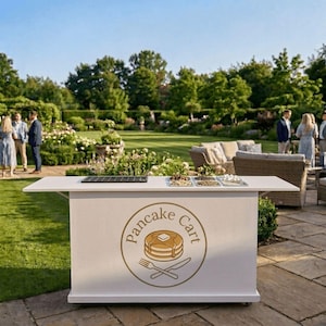 May include: A white pancake cart with a gold logo and text that reads "Pancake Cart". The cart has a countertop with toppings and a griddle. The cart is outdoors with people in the background.