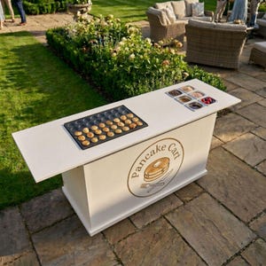 May include: A white pancake cart with a black griddle filled with small pancakes. The cart has a logo that says "Pancake Cart" and includes a stack of pancakes with a fork and knife. Various toppings are in small containers.