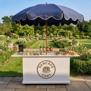 May include: A white pancake cart with a navy blue scalloped umbrella. The cart has a gold-colored logo that says "PANCAKE CART". The cart is stocked with bottles, jars, and bowls of toppings. The cart is set up outdoors in a garden.