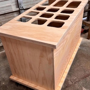 Peut inclure: Un coffre de rangement en bois clair de forme rectangulaire. Le dessus du coffre comporte une série de découpes carrées. Le coffre est en contreplaqué et repose sur une base.