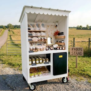 May include: A white wooden mobile farm stand with a scalloped roof, featuring multiple shelves stocked with various packaged goods, jars, and a small black refrigerator. A wooden sign nearby displays the text FARM STAND in black lettering.