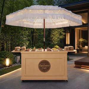 May include: A light wood charcuterie cart featuring a circular logo with the text Charcuterie Cart, topped with a large white fringed umbrella illuminated by integrated string lights and multiple trays of food arranged on the counter surface.