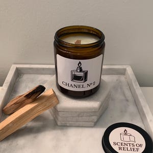 K&ouml;nnte beinhalten: Ein braunes Glas-Kerzenglas mit einem wei&szlig;en Etikett mit der Aufschrift "CHANEL N&deg;2" und einer Parf&uuml;mflaschen-Grafik. Die Kerze steht auf einem Marmortablett mit Palo-Santo-St&auml;bchen und einem schwarzen Deckel mit "SCENTS OF RELIEF".