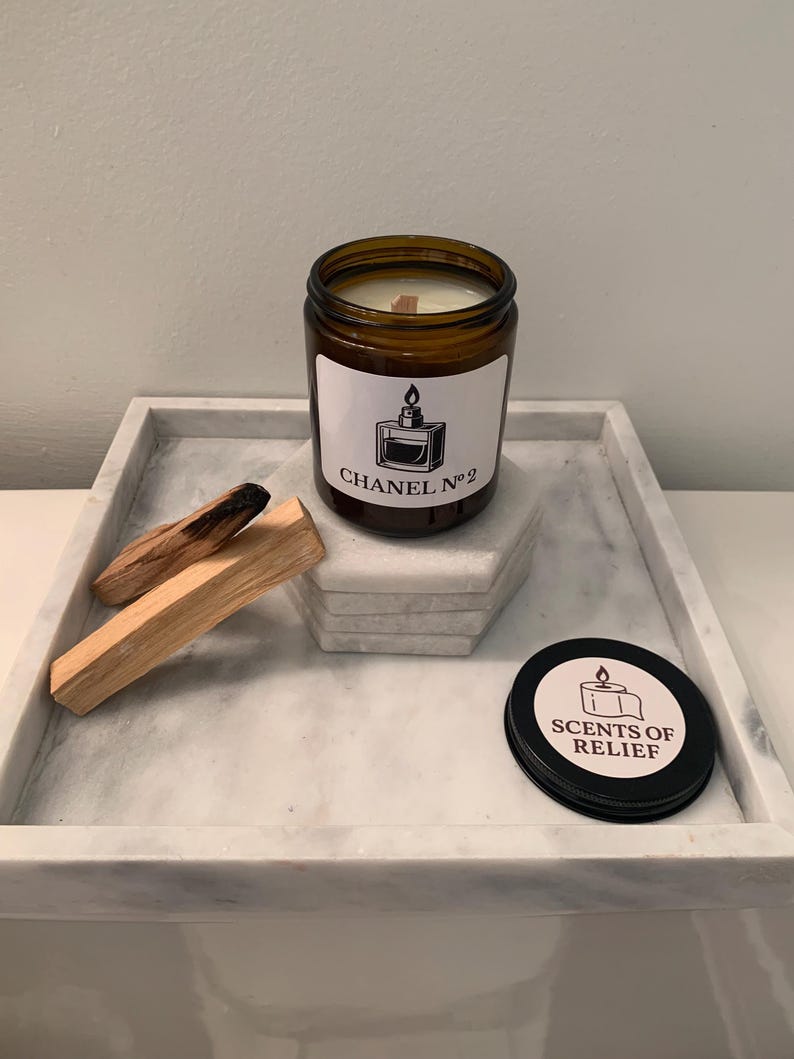 K&ouml;nnte beinhalten: Ein braunes Glas-Kerzenglas mit einem wei&szlig;en Etikett mit der Aufschrift "CHANEL N&deg;2" und einer Parf&uuml;mflaschen-Grafik. Die Kerze steht auf einem Marmortablett mit Palo-Santo-St&auml;bchen und einem schwarzen Deckel mit "SCENTS OF RELIEF".