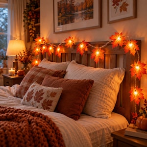 May include: A string of LED lights featuring artificial orange and red maple leaves draped across a wooden headboard. The lights emit a warm glow, illuminating the bed which is covered with a chunky knit blanket and several decorative pillows.