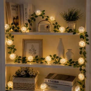 May include: A string of artificial green vines featuring illuminated white rose-shaped LED lights. The glowing floral garland is draped across multiple shelves, winding around books, a small radio, vases, and a potted plant, emitting a warm light.
