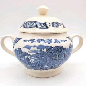 May include: Cream-colored ceramic sugar bowl with a lid, featuring blue pastoral scenes and floral patterns. The bowl has two handles and a knob on the lid. The design is reminiscent of vintage tableware.