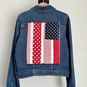May include: A blue denim jacket with a patchwork American flag design on the back. The flag features red and white stripes, white polka dots, and a blue field with white stars. The jacket is hanging on a wooden hanger.