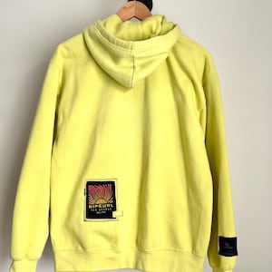 May include: A bright yellow hoodie with a drawstring hood and long sleeves. The back of the hoodie features a rectangular patch with the text "RIP CURL THE SEARCH" in a stylized design. A small black patch is on the left sleeve.