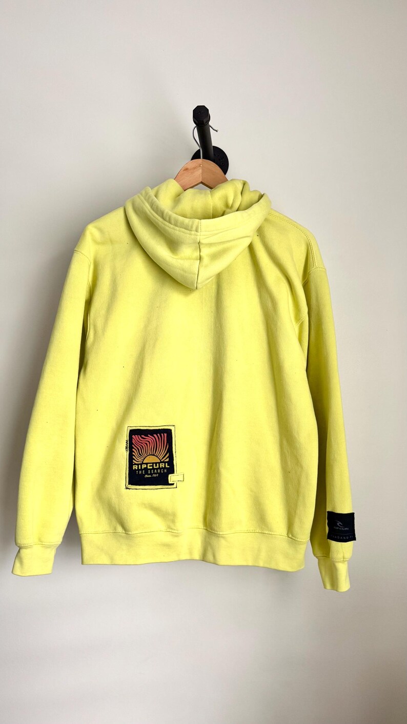 May include: A bright yellow hoodie with a drawstring hood and long sleeves. The back of the hoodie features a rectangular patch with the text "RIP CURL THE SEARCH" in a stylized design. A small black patch is on the left sleeve.