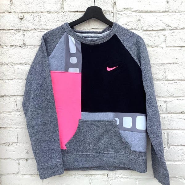 May include: A gray, black, and pink sweatshirt with a Nike logo. The sweatshirt has a color block design with a kangaroo pocket. The sleeves and pocket are gray, with a black panel and pink panel on the front.