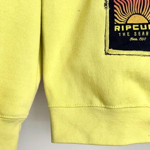 May include: A yellow sweatshirt with a black and orange graphic on the front. The graphic features a sun design with the text "RIPCURL THE SEARCH Since 1969". The sweatshirt has ribbed cuffs and hem.
