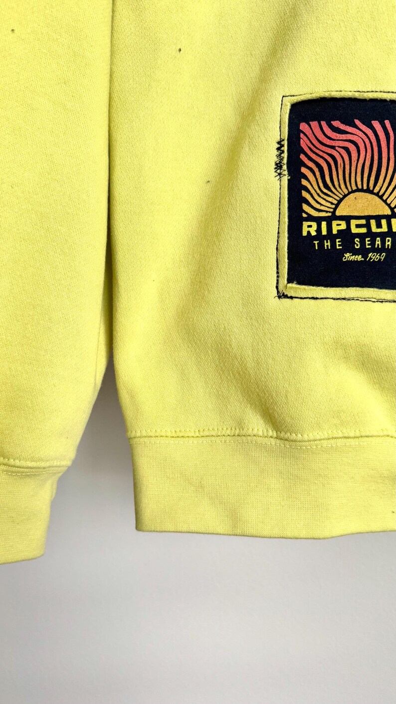 May include: A yellow sweatshirt with a black and orange graphic on the front. The graphic features a sun design with the text "RIPCURL THE SEARCH Since 1969". The sweatshirt has ribbed cuffs and hem.