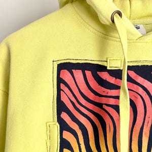 May include: A yellow hoodie with a graphic design featuring a stylized sun in shades of orange and yellow against a dark blue background. The hoodie has a drawstring and a hood. The design is framed with a black stitched border.