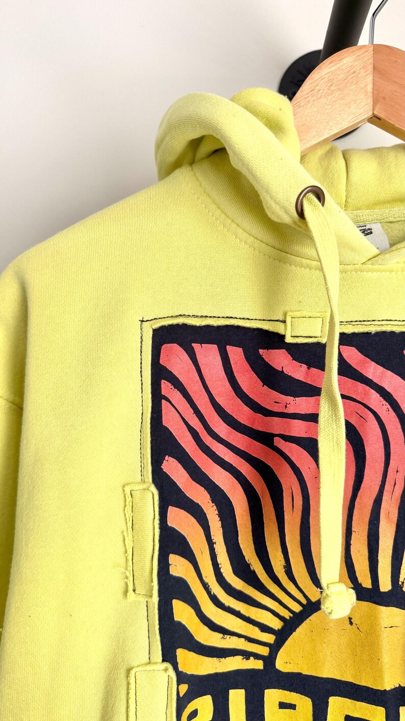 May include: A yellow hoodie with a graphic design featuring a stylized sun in shades of orange and yellow against a dark blue background. The hoodie has a drawstring and a hood. The design is framed with a black stitched border.
