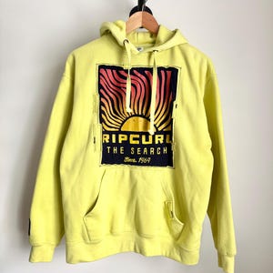 May include: A yellow hoodie with a front pocket and a hood. The front features a graphic of a sunset with the text "RIP CURL THE SEARCH Since 1969" in black and red. The hoodie is hanging on a wooden hanger.