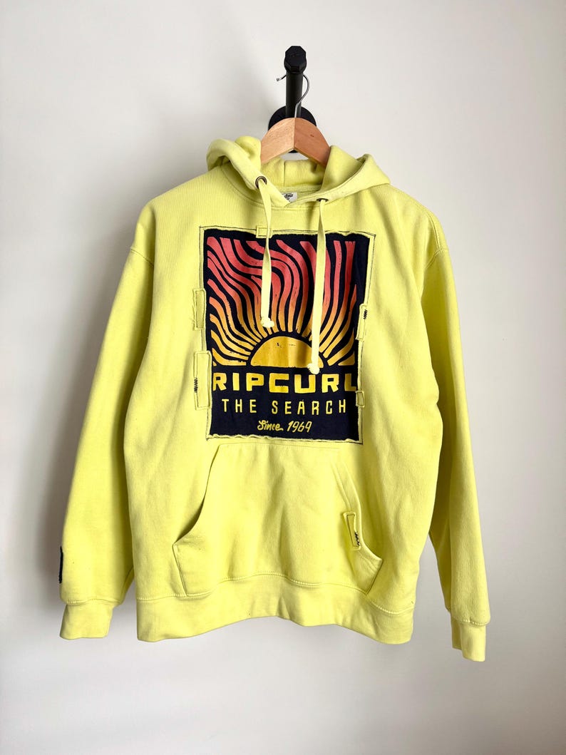 May include: A yellow hoodie with a front pocket and a hood. The front features a graphic of a sunset with the text "RIP CURL THE SEARCH Since 1969" in black and red. The hoodie is hanging on a wooden hanger.