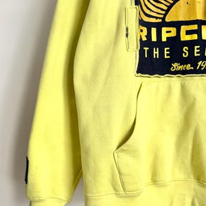 May include: A yellow hoodie with a front pocket and a drawstring hood. The hoodie features a large graphic on the upper chest with a sun design in orange, yellow, and black, and the text "RIPCURL THE SEARCH Since 1969".