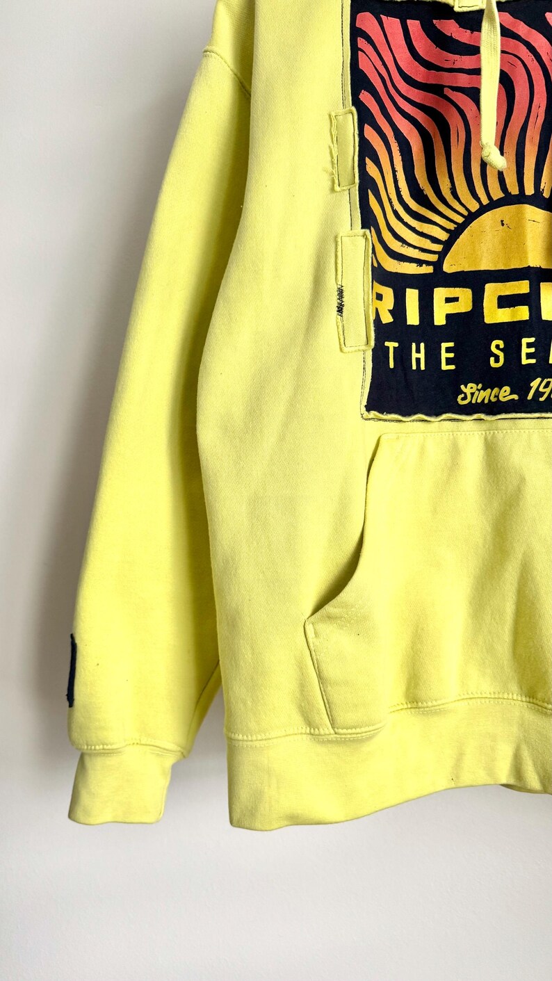 May include: A yellow hoodie with a front pocket and a drawstring hood. The hoodie features a large graphic on the upper chest with a sun design in orange, yellow, and black, and the text "RIPCURL THE SEARCH Since 1969".