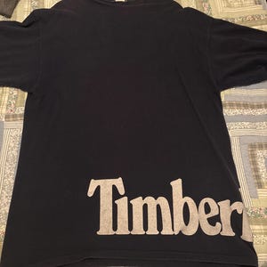 Pode incluir: Uma camiseta azul marinho com a palavra "Timber" impressa em bege claro na parte inferior da frente. A camiseta está pendurada em um cabide branco.
