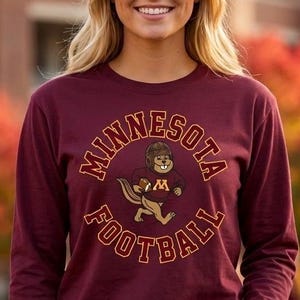 Puede incluir: Una camiseta de manga larga color granate con un gráfico de una ardilla listada de dibujos animados que lleva un casco de fútbol americano de cuero vintage y un suéter granate con una M dorada, rodeada por el texto MINNESOTA FOOTBALL.