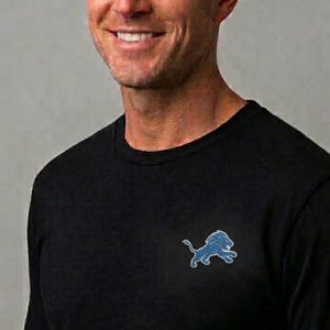 May include: A black long-sleeve shirt featuring a small, light blue embroidered lion logo on the left chest. The shirt has a classic crew neckline and a simple, solid black fabric construction, providing a clean and understated appearance.