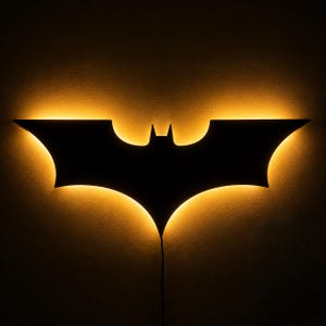 Puede incluir: Un aplique de pared con la forma del símbolo del murciélago de Batman. La silueta negra sólida está retroiluminada con una luz ámbar cálida y brillante, creando un suave efecto de halo contra la pared que resalta el icónico diseño de las alas.