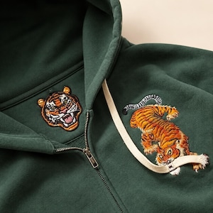 Op de afbeelding: Een donkergroene hoodie met rits en twee geborduurde tijgerontwerpen. Eén patch toont een tijgerkop bij de kraag, terwijl een tweede tijger in zijn geheel op de borst is geborduurd en gedeeltelijk het crèmekleurige trekkoord overlapt.