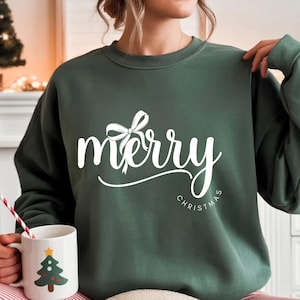 Könnte beinhalten: Ein dunkelgrünes Sweatshirt mit den Wörtern "merry" und "Christmas" in weißer Schrift. Eine weiße Schleifengrafik befindet sich über dem Wort "merry". Eine weiße Tasse mit einem Weihnachtsbaum-Design wird in der Hand gehalten.