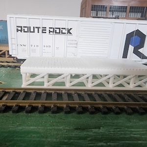 May include: A white model train loading dock with a wooden plank design, supported by a white truss structure. The words "ROUTE ROCK" are printed on a white train car in the background. Model train tracks are in the foreground.