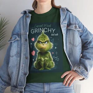 Op de afbeelding: Groen T-shirt met een chagrijnige Grinch-personage en de tekst "Current Mood Grinchy but cute". Het ontwerp bevat roze harten en sterren. De persoon draagt een blauw spijkerjasje.