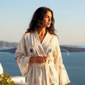 May include: A long-sleeved, off-white robe featuring a repeating embroidered evil eye pattern in shades of blue and brown. The garment has a wrap-around waist tie, a V-neckline, and frayed edges along the sleeve cuffs and the bottom hemline.