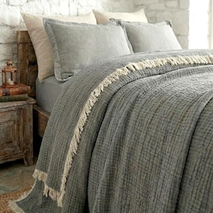 May include: A grey bedspread with a textured surface and a cream-coloured fringe. The bed is made up with grey pillows and a matching bed sheet. The room has a rustic aesthetic with a white brick wall and wooden furniture.