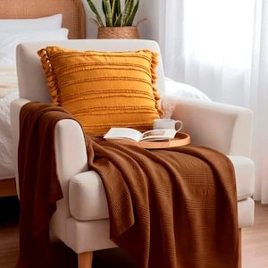 May include: A cream-colored armchair featuring a mustard yellow square throw pillow with fringe tassels and horizontal textured stripes. A dark brown waffle-knit throw blanket is draped over the seat, holding a wooden tray with an open book.
