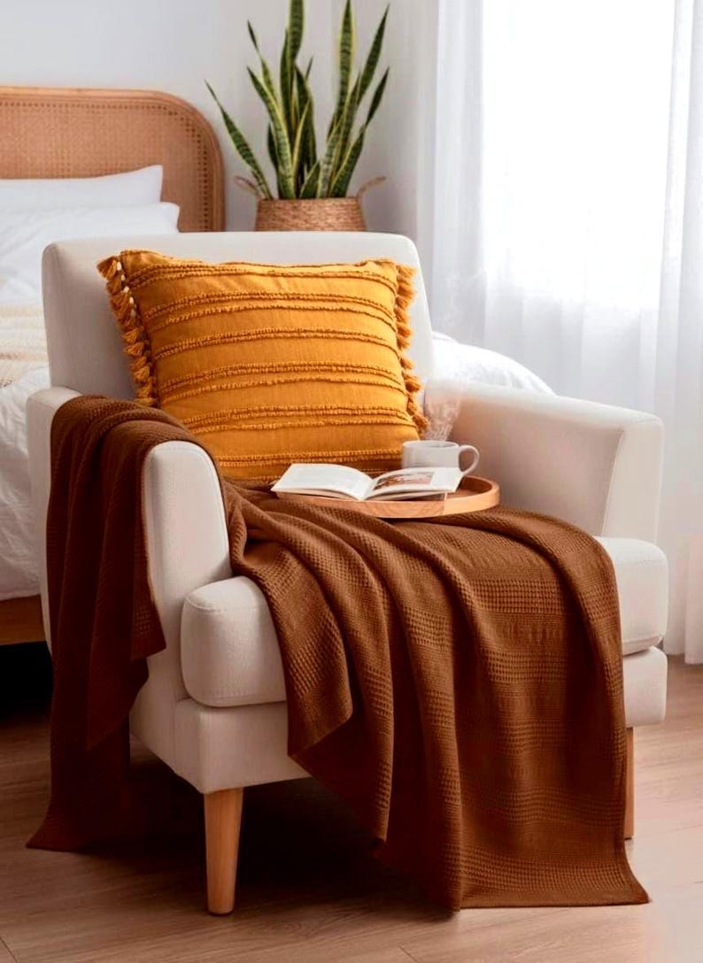 May include: A cream-colored armchair featuring a mustard yellow square throw pillow with fringe tassels and horizontal textured stripes. A dark brown waffle-knit throw blanket is draped over the seat, holding a wooden tray with an open book.