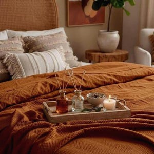 May include: A rectangular wooden tray sits on a textured rust-colored waffle-weave blanket. The tray holds two glass reed diffuser bottles with wooden sticks, a small ceramic bowl containing dried botanicals, and a lit white candle in a glass.