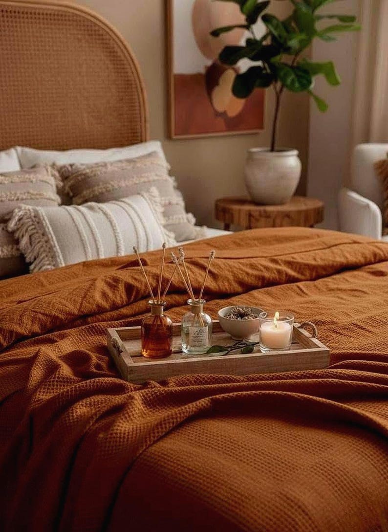 May include: A rectangular wooden tray sits on a textured rust-colored waffle-weave blanket. The tray holds two glass reed diffuser bottles with wooden sticks, a small ceramic bowl containing dried botanicals, and a lit white candle in a glass.