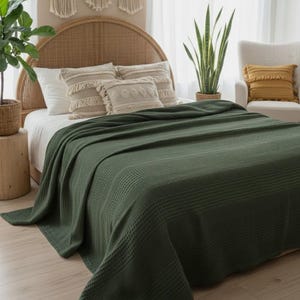 May include: A dark olive green waffle-weave blanket draped over a bed. The blanket features a textured grid pattern across its entire surface, providing a uniform appearance. It is positioned to cover the mattress, extending down to the floor.